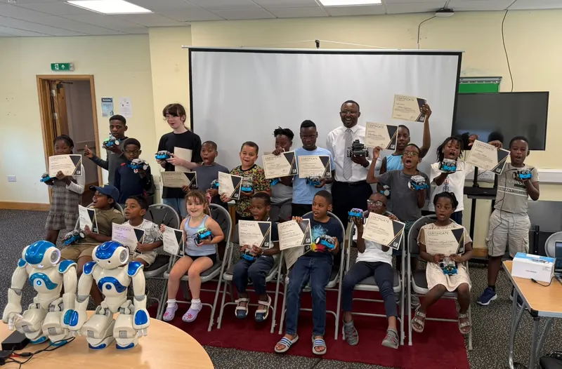 A group of children proudly holding their certificates and robots at a Kids in Tech award ceremony, with two humanoid robots in the foreground.