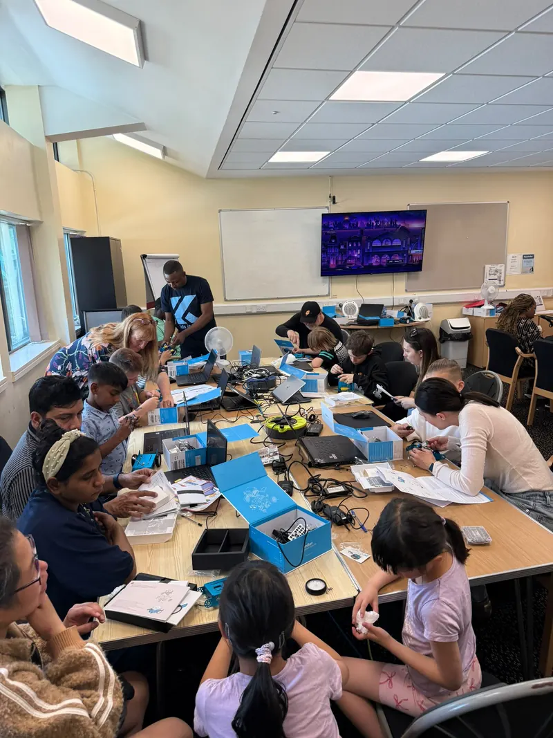 Children and parents around a long table building robot kits with laptops during a Kids in Tech workshop.
